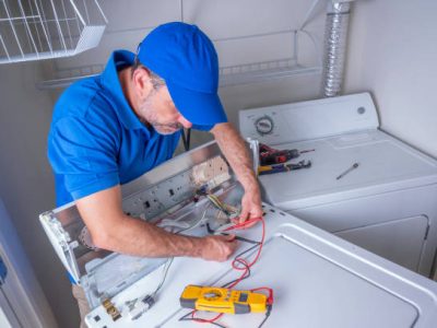 Appliance service technician in a blue uniform testing circuits on a washing machine with a volt meter