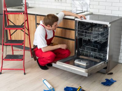 Technician or worker in uniform installs dishwasher into the kitchen furniture. Repairman wear worker suit repairing maintenance of dishwasher. Master in protective gloves fix dishwasher.